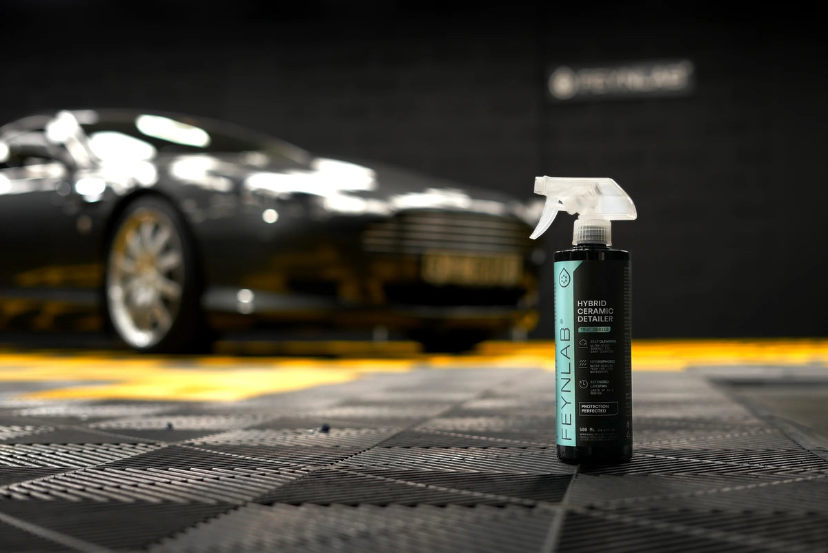 Ciemny Aston Martin w studiu detailingu z logo Feynlab na ścianie, na pierwszym planie butelka Feynlab Hybrid Ceramic Detailer
